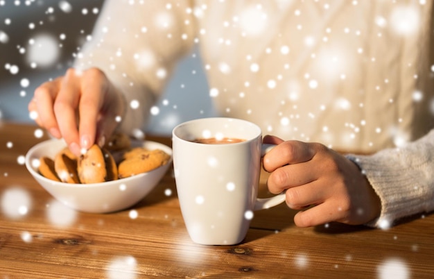 inverno, cibo e bevande concetto - primo piano di donna con biscotti di avena e tazza di cioccolata calda seduta al tavolo di legno a casa