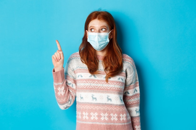 Inverno, covid-19 e concetto di quarantena. Curiosa ragazza rossa in maschera medica che raccoglie prodotto, guardando e indicando l'angolo in alto a sinistra promozionale, sfondo blu.
