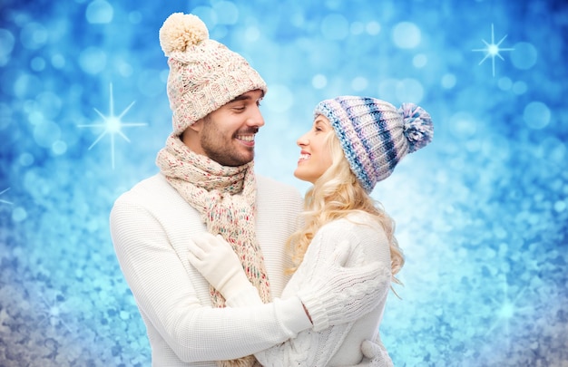 inverno, moda, coppia, natale e concetto di persone - uomo e donna sorridenti con cappelli e sciarpa che si abbracciano su sfondo blu glitter e luci natalizie