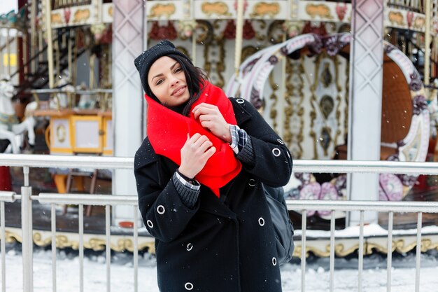 Inverno, natale, stile di vita, vacanze e concetto di moda - Donna sorridente abbastanza giovane in cappotto nero e sciarpa rossa che posano nella città d'inverno. attraente bruna sfondo della giostra francese