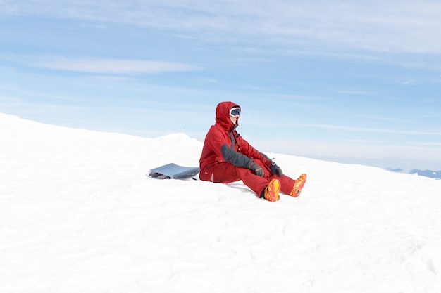 Inverno, tempo libero, sport e concetto di persone: lo snowboarder siede in alto sulle montagne sul bordo del pendio e guarda in lontananza.