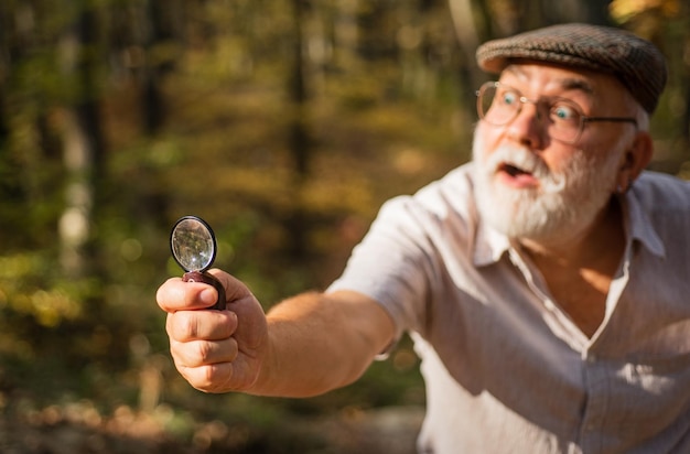 Investigazione privata Zoom Concetto di curiosità Detective esigente nella foresta Esplora la natura Pensionato con lente d'ingrandimento esplorando la foresta messa a fuoco selettiva Vecchio scienziato Nonno barbuto nella foresta