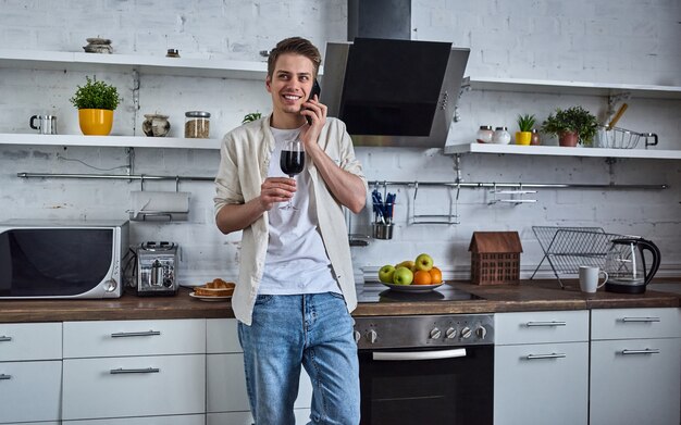 Invitare gli amici a cena. Bel giovane in abbigliamento casual che beve vino e parla al telefono mentre sta in piedi in cucina a casa