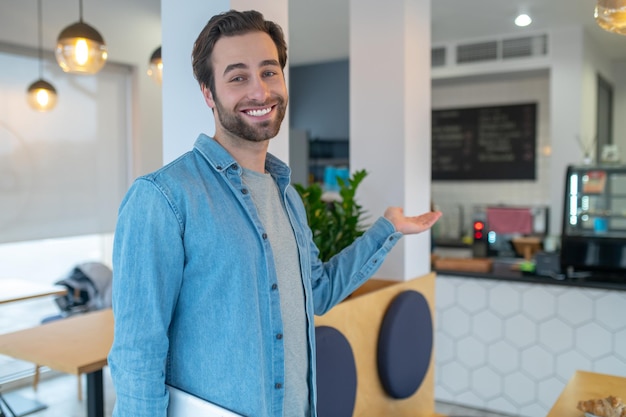 Invito. Attraente giovane uomo barbuto con un sorriso a trentadue denti che guarda affabilmente la fotocamera con un gesto che invita nella caffetteria