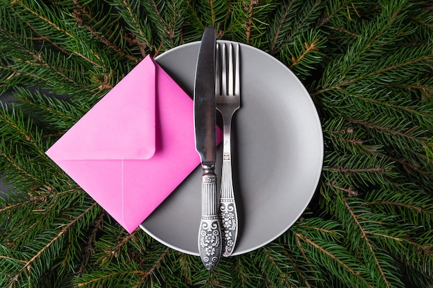 Invito per il concetto di cena festiva. Busta rosa, forchetta, coltello e piatto su sfondo di rami di abete. Foto di alta qualità