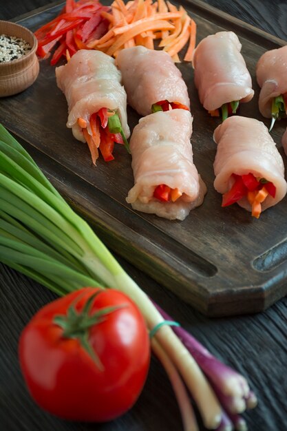 Involtini fatti in casa con petto di pollo fresco alle erbe, fette di carote, peperone su un tagliere scuro. L'equilibrio di un'alimentazione sana. Cucinando. Sfondo di legno scuro