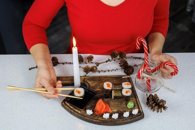 Involtini giapponesi con pesce rosso su un tagliere di legno Una ragazza con un maglione rosso mangia sushi con le bacchette Decorazione della tavola con candele e dolci Concetto natalizio