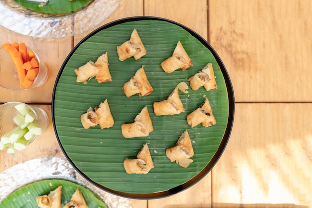 Involtini primavera fritti con verdure e vermicelli su piatto nero circolare con foglia di banana su un tavolo di legno all'aperto