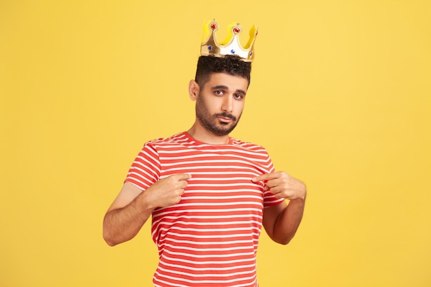 io sono il re. Ambizioso uomo barbuto che indossa una corona d'oro e si indica, guardando con arroganza, dichiarando la sua autorità, status privilegiato superiore. Studio indoor girato isolato su sfondo giallo