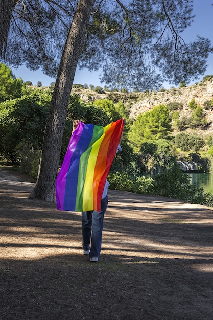 Irriconoscibile donna lesbica di mezza età che tiene la bandiera arcobaleno gay sul lago all'aperto. Concetto di libertà