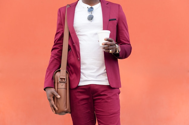 Irriconoscibile uomo d'affari nero elegante vestito in abito con valigetta tiene il caffè in mano davanti a sfondo rosso, copia spazio per il testo