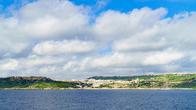 Isola di Gozo, Malta. Cielo nuvoloso blu in primavera, erba verde giovane e vista su Mgarr e sul forte Chambray. Panorama vista mare. Sfondo orizzontale lungo, concetto di vacanza da cartolina di viaggio.