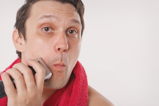 Isolato: un uomo si rade la barba. il ragazzo si pulisce la barba con un rasoio elettrico. trattamenti mattutini in bagno. asciugamano rosso intorno al collo. copia spazio