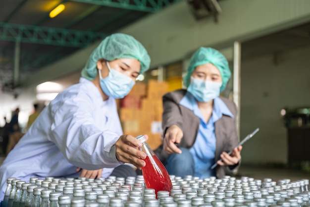Ispettore asiatico supervisore donna con assistente dipendente che indossa una tuta sterile che controlla il prodotto in stock di confezioni di bevande alla frutta in magazzino presso l'impianto di lavorazione
