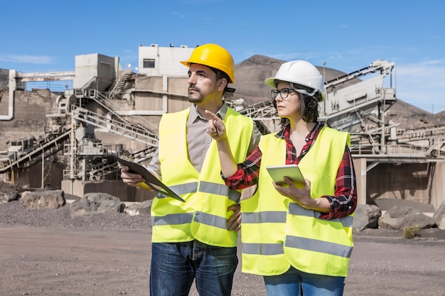 Ispettore femminile professionale serio con la compressa che indica via chiedendo parere dell'ingegnere maschio con appunti sull'oggetto di ingegneria mentre visita il cantiere industriale