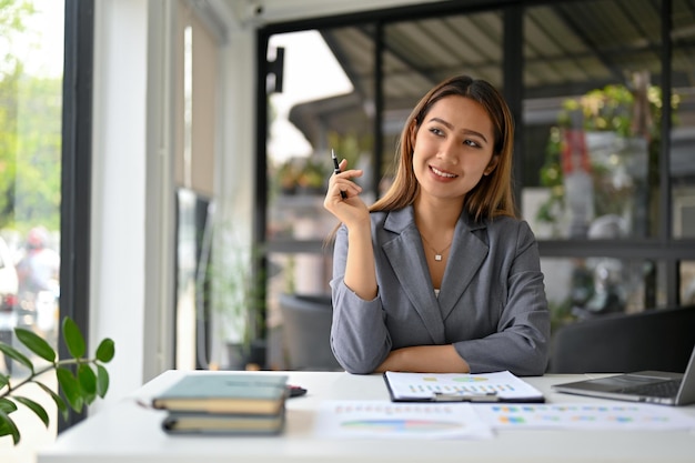 Ispirata donna d'affari asiatica che pensa e sogna ad occhi aperti sul suo percorso professionale alla sua scrivania