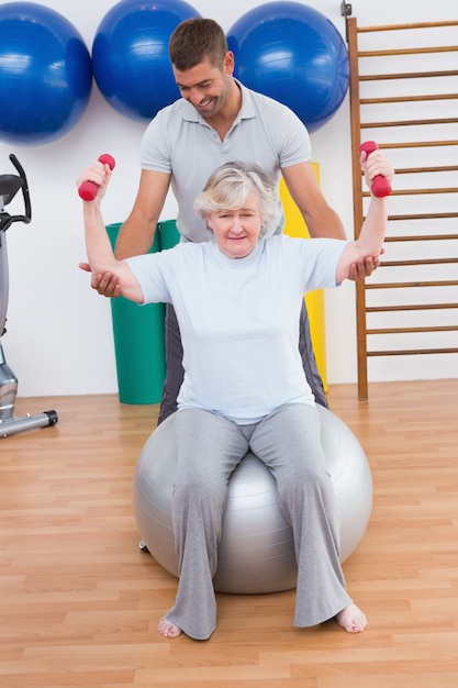 Istruttore che aiuta i dumbbells dell&#39;elevatore della donna senior sulla palla di esercizio