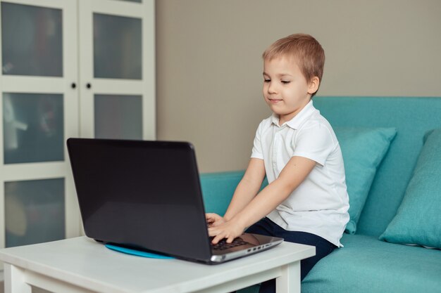 istruzione a distanza domiciliare dei bambini durante la quarantena. felice bambino in polo bianco a fare i compiti utilizzando il computer portatile seduto sul divano