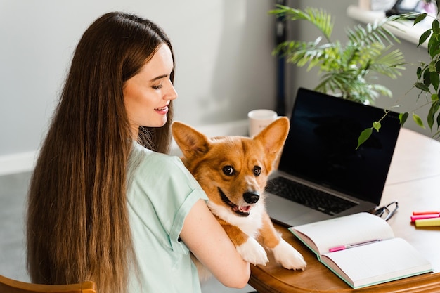 Istruzione a distanza Studentessa con laptop studia online con il suo adorabile cane Corgi a casa Abbraccio di giovane donna Welsh Corgi Pembroke e prendere appunti sul taccuino Stile di vita con animale domestico