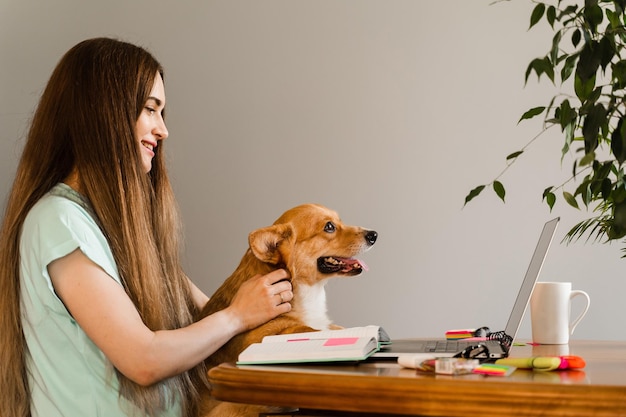 Istruzione a distanza Studentessa con laptop studia online con il suo adorabile cane Corgi a casa Abbraccio di giovane donna Welsh Corgi Pembroke e prendere appunti sul taccuino Stile di vita con animale domestico