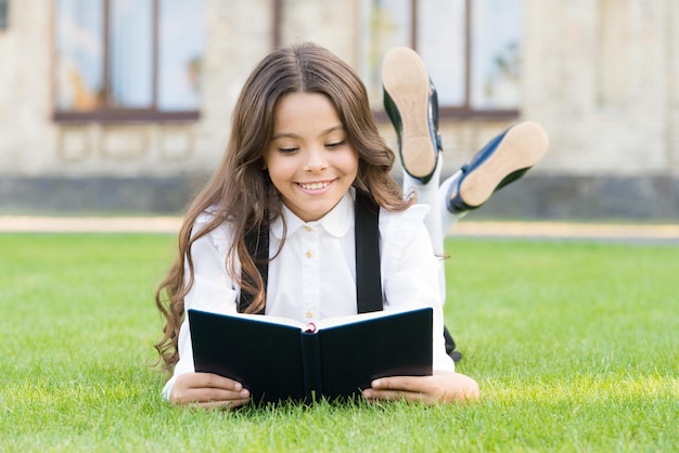 Istruzione di base Adorabile bambina impara a leggere Studentessa uniforme scolastica sdraiata sul prato con il libro preferito Concetto di studio Lettura extracurricolare Carino bambino piccolo libro di lettura all'aperto