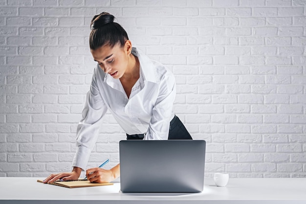 Istruzione online e concetto di studio su Internet con una giovane donna graziosa in camicia bianca che prende appunti in un blocco note appoggiato su un tavolo di legno bianco con laptop e tazza di caffè su uno sfondo di muro di mattoni chiari