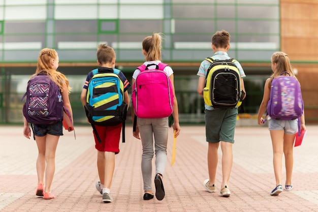 istruzione primaria, amicizia, infanzia e concetto di persone - gruppo di studenti delle scuole elementari felici con zaini che camminano all'aperto dalla parte posteriore