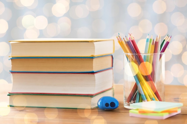 istruzione, scuola, creatività e concetto di oggetto - primo piano di pastelli o matite colorate con libri, adesivi e temperino su tavolo di legno durante le vacanze luci sfondo