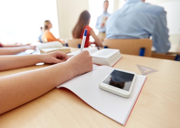 istruzione, scuola superiore, apprendimento, tecnologia e concetto di persone - primo piano delle mani degli studenti che scrivono su notebook e smartphone sulla scrivania