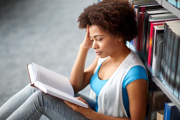 istruzione, scuola superiore, università, apprendimento e concetto di persone - ragazza studentessa afroamericana che legge libro seduto sul pavimento in biblioteca