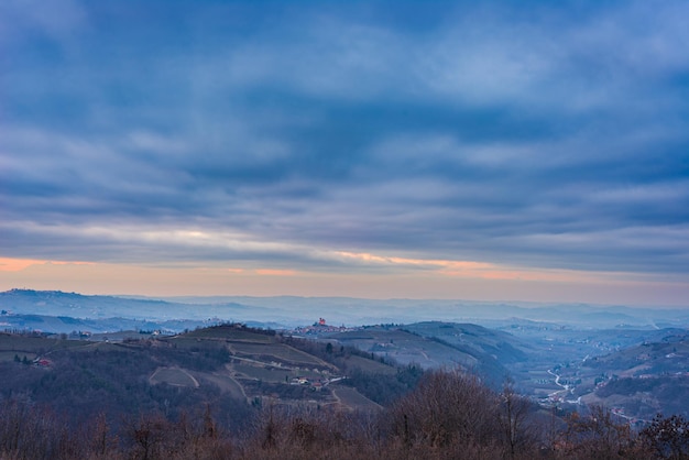 Italia Piemonte: panoramica neve invernale vista vigneti paesaggio unico al tramonto, castello medievale e villaggio in cima a una collina, le Alpi sullo sfondo cielo drammatico