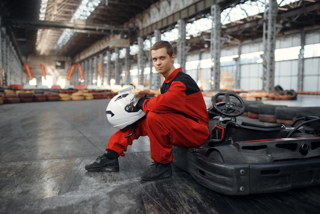 Kart racer in uniforme rossa posa con hemet in mano, kart auto sport indoor. Gara di velocità su pista di go-kart ravvicinata con barriera per pneumatici. Gara di veicoli veloci, hobby ad alta adrenalina