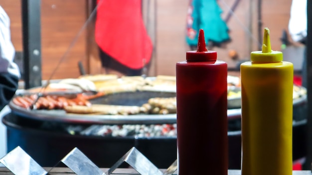 Ketchup e maionese in tubi di plastica vicino a una griglia di strada in primo piano in primo piano con messa a fuoco selettiva. Barbecue, festival del cibo di strada. Due lattine di sughi di carne. Contenitore rosso e giallo.