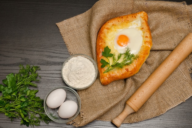 Khachapuri con le uova su tela di sacco, farina, uova e prezzemolo sul tavolo nero