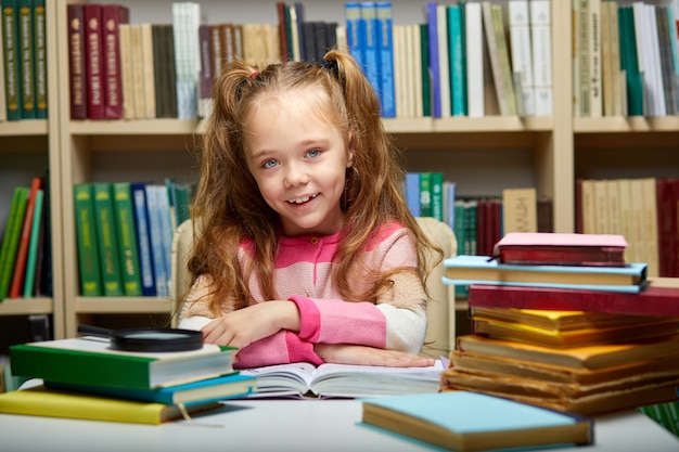 Kid girl si siede con i libri a tavola in biblioteca, bambino in libreria, circondato da libri colorati per la scuola, guarda sorridente della fotocamera