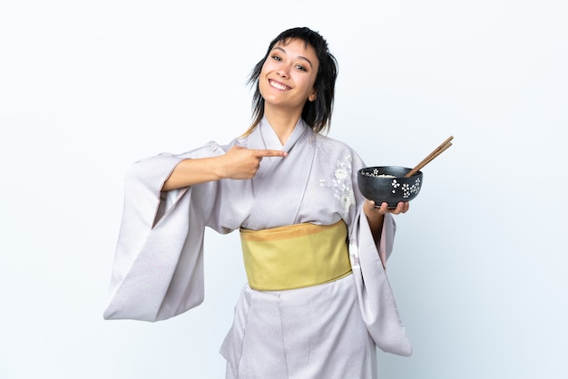 Kimono d'uso della giovane donna che tiene una ciotola di tagliatelle sopra bianco isolato e che lo indica