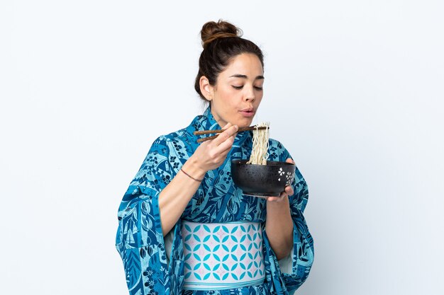 Kimono da portare della donna sopra la parete bianca isolata che tiene una ciotola di tagliatelle con la sabbia della bacchette che lo soffia perché sono calde