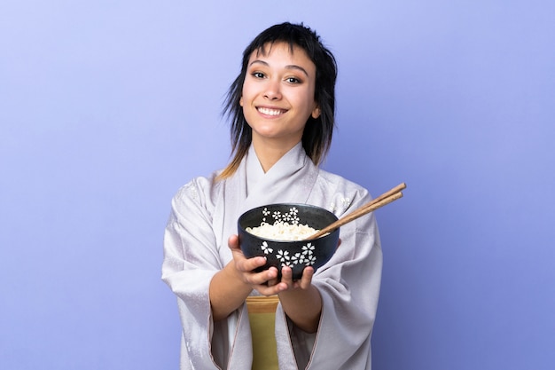 Kimono da portare della giovane donna sopra la parete blu con l'espressione felice mentre tenendo una ciotola di tagliatelle con le bacchette