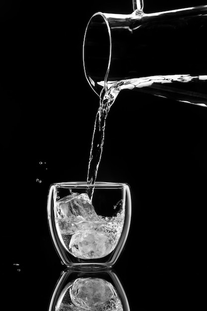 L'acqua versa in un bicchiere con ghiaccio da un decanter su sfondo nero