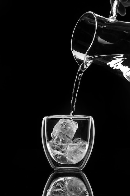 L'acqua versa in un bicchiere con ghiaccio da un decanter su sfondo nero