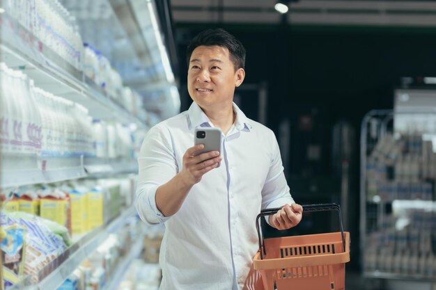 L'acquirente asiatico cammina intorno al supermercato con un carrello guardando la navigazione dello smartphone