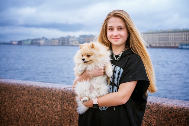 L'adolescente allegro e felice tiene e abbraccia lo spitz di Pomerania sull'argine in città in una giornata nuvolosa...