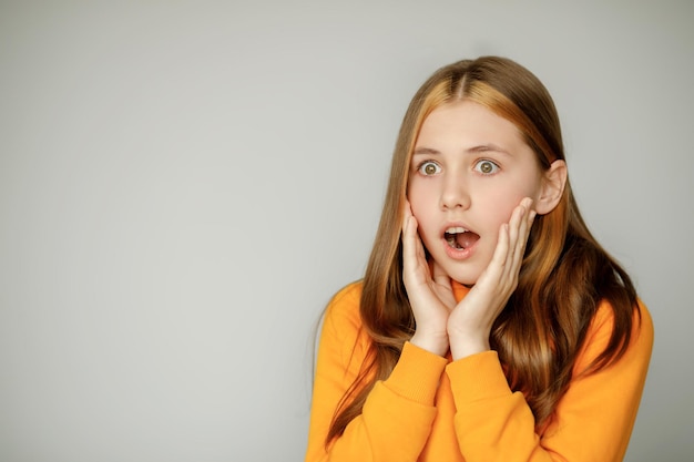 L'adolescente con un maglione arancione con gli occhi spalancati è molto sorpresa di abbracciarsi il viso con la mano