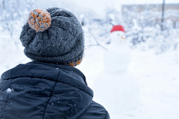 L'adolescente dell'inverno in un rivestimento e un cappello scolpisce un pupazzo di neve su un fondo dell'inverno con la neve di caduta nel concetto congelato del giorno