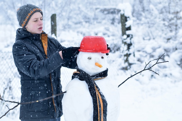 L'adolescente dell'inverno in un rivestimento e un cappello scolpisce un pupazzo di neve su un fondo dell'inverno con la neve di caduta nel concetto congelato del giorno