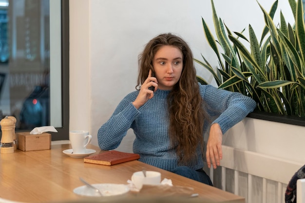 L'adolescente sta parlando al telefono nella caffetteriaLa ragazza con lunghi capelli castani si siede nella caffetteria con una tazza di tè o caffè e un taccuino
