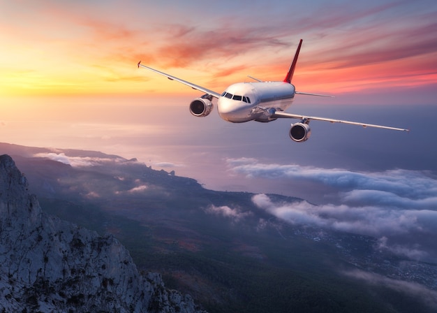 L'aereo sta volando sopra le nuvole basse al tramonto. Paesaggio con aereo passeggeri, montagne, mare e cielo arancione con nuvole rosse in estate. Aerei passeggeri. Viaggi d'affari in Europa. Aereo commerciale