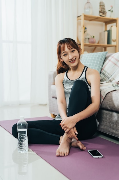 l'affascinante ragazza asiatica verticale in abbigliamento sportivo sorride alla telecamera mentre si riposa sul tappetino da yoga con il suo telefono e un'acqua in bottiglia dopo l'esercizio a casa.