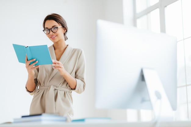 L'allegra regista femminile tiene l'organizzatore personale guarda attraverso i piani per il giorno indossa occhiali in costume formale beige posa vicino al dektop con un grande monitor sfondo bianco verifica le informazioni registrate