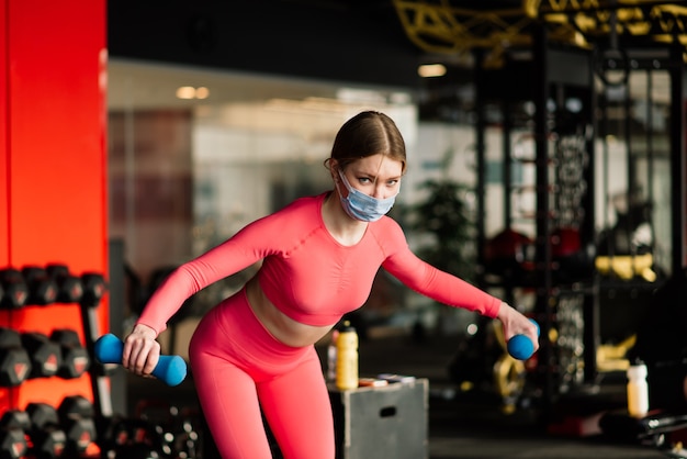 L'allenatore di fitness dell'atleta femminile che indossa la maschera protettiva medica bianca fa esercizi con manubri, invita a fare lezione di fitness in palestra vuota.